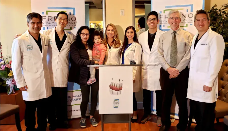 Surgeons in white coats standing with staff and community members, one holding a small child, in front of Fresno Oral Maxillofacial Surgery banners